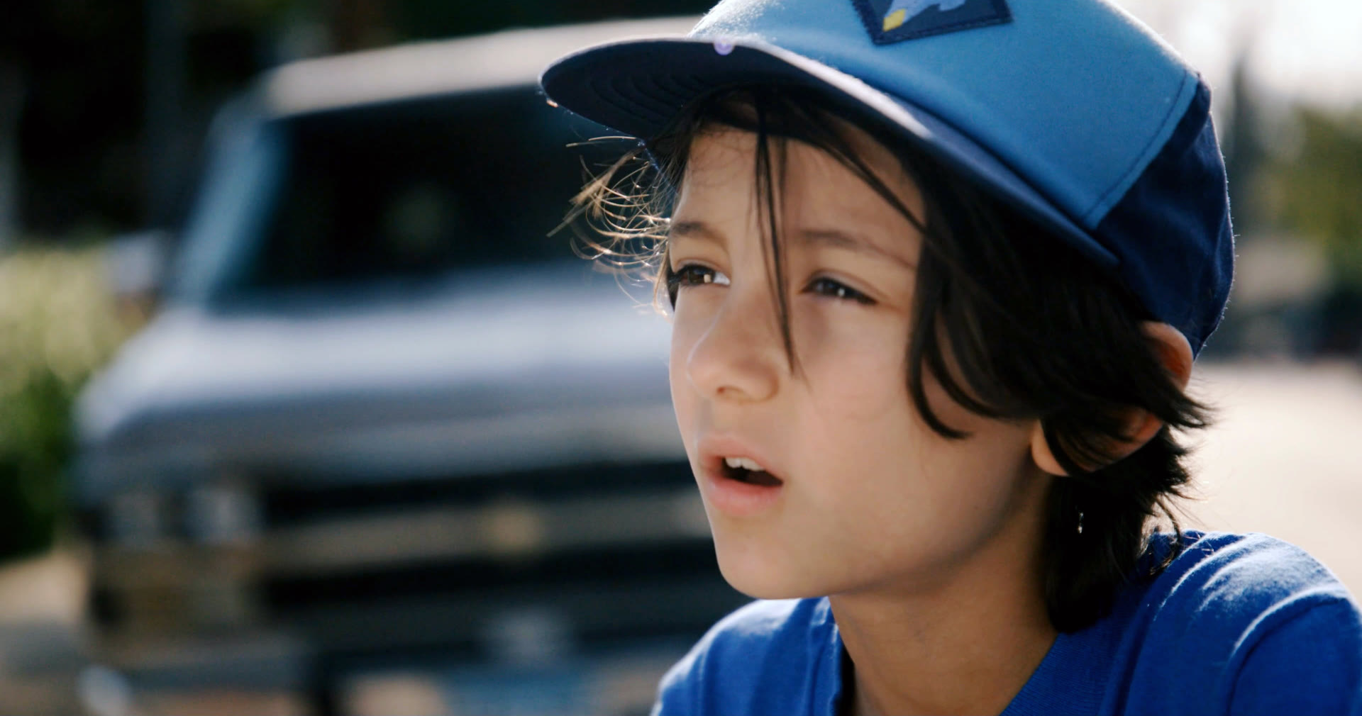 Close-up of a boy in a blue cap and tee looking off camera, a truck grille soft behind him.
